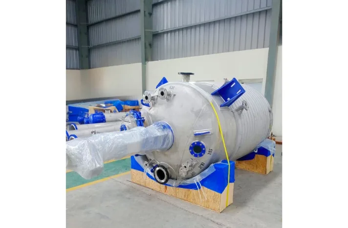 It is a stainless steel high-pressure reactor with a flange and limpet coil located on a wooden pallet in a factory.